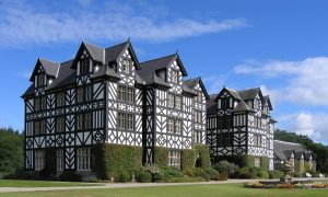 An 1840's mansion with concrete cladding designed to replicate the black and white timber-framed architecture of Montgomeryshire farmhouses. Set back slightly on green grass, with a long driveway. The sky is bright with just one or two clouds.