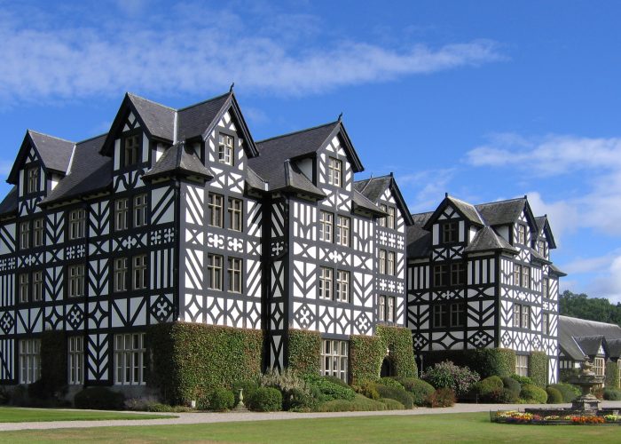 An 1840's mansion with concrete cladding designed to replicate the black and white timber-framed architecture of Montgomeryshire farmhouses. Set back slightly on green grass, with a long driveway. The sky is bright with just one or two clouds.