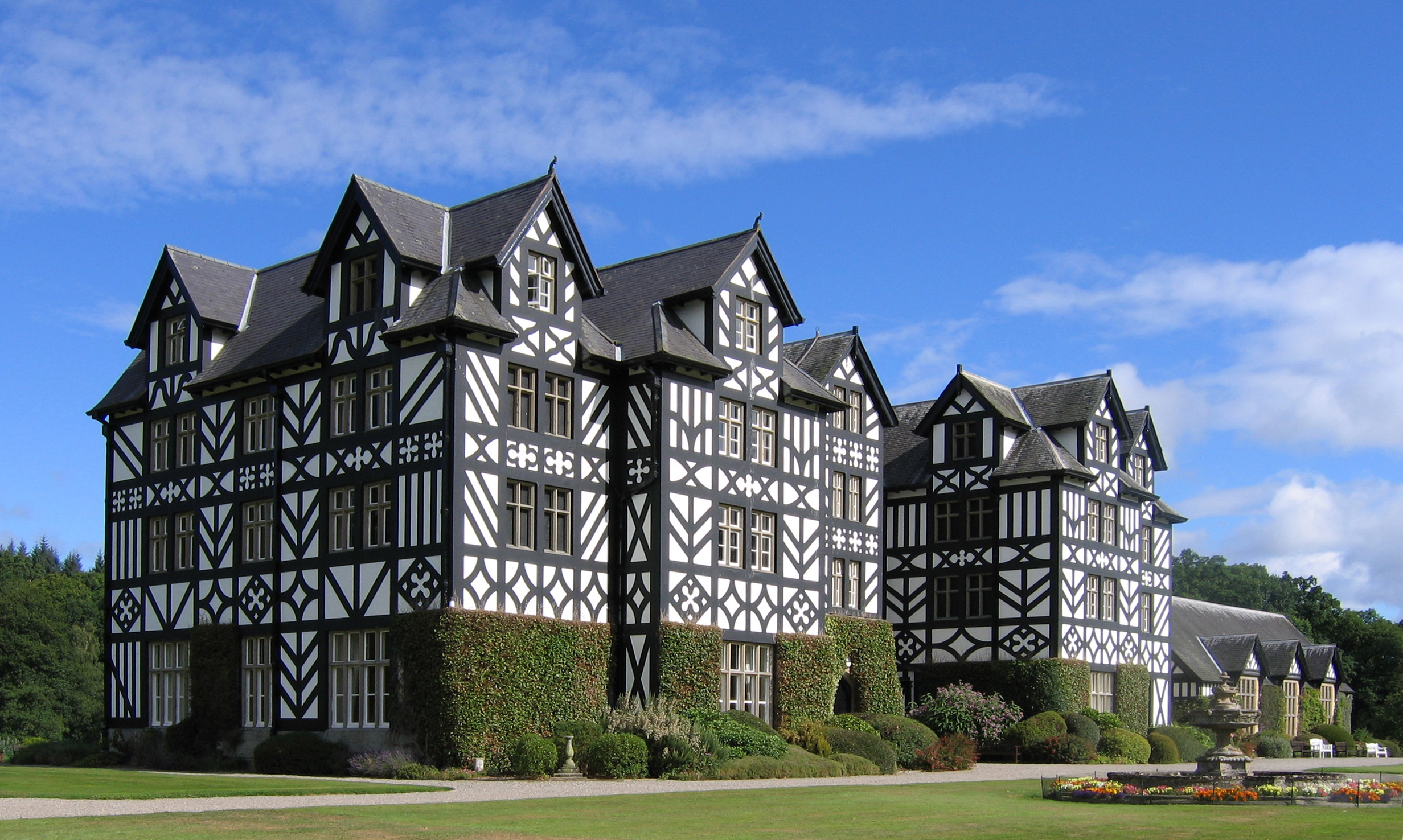 An 1840's mansion with concrete cladding, designed to replicate the black-and-white timber-framed architecture of Montgomeryshire farmhouses. Set in green grass grounds with a fountain to the right of the image. A bright sky with a few clouds.